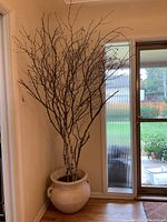 Full view of the decorative artificial tree with thin branches in the white pot placed near glass door and window showing overall height.