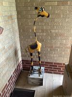 Front-left angled view of the large metal giraffe sculpture mounted on a gray wooden box stand, showing yellow and brown paint finish and metal flowers on head.
