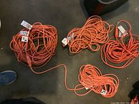 Top-down view of four coiled bright orange heavy duty extension cords on concrete floor, each cord tagged with labels.