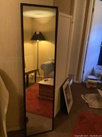 Full-length black framed mirror leaning against a wall showing reflection of floor lamp, furniture, and room interior.