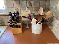 Knife block filled with various kitchen knives next to a utensil crock holding wooden and plastic utensils on kitchen counter.