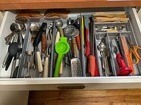 Top-down image of kitchen utensil drawer with metal mesh drawer dividers separating various utensils like wooden spoons, peelers, graters, ladles, scissors, and measuring spoons.