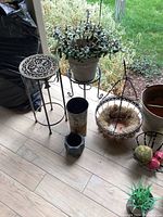 Photo shows small and medium black metal plant stands, ceramic glazed planter with artificial succulents, and several other decorative pots and baskets on tiled floor.