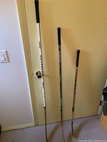 Photo of three hockey sticks standing against a door: one tall white Sher-Wood D500, one medium Easton Synergy, and one short natural wood Sher-Wood stick.