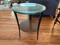 Full view of the modern round side table showing the thick glass top, metal frame, and lower shelf on hardwood floor.