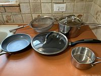 Photo showing the full assortment of pots, pans, strainer, and glass lid on countertop.