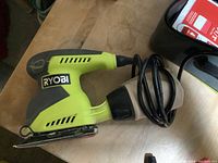 Close-up image of Ryobi electric hand sander with dust bag attached, showing green and black plastic body with cord coiled on side.