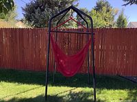 Outdoor red swing chair hanging from black metal stand on grassy lawn against wooden fence background.