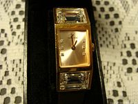 Front view of Swarovski watch showing gold tone case, champagne dial with rhinestone hour markers, and large rectangular clear rhinestones above and below dial.