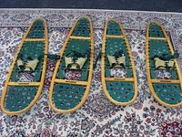 Top-down view of four snow shoes on an oriental style carpet, showing two larger and two smaller pairs with wooden frames and green deck surfaces with bindings.