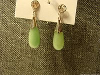 Front view showing green jade teardrop pendants hanging from gold-tone engraved character posts on fabric background.