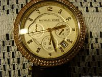 Close-up image of watch face showing gold tone dial, rhinestone hour markers and bezel, chronograph subdials, and date window.