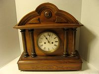 Front view of the antique wooden mantel clock showing carved wood case, clock face with Roman numerals, and decorative columns.