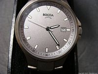 Close-up of Boccia watch face showing clean matte gray dial, date window at 3, luminous minute/hour hands, and second hand. Titanium case visible with minimal wear.