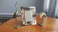 Wide shot showing both brass bird figurines and the decorated photo frame on a wooden table indoors.