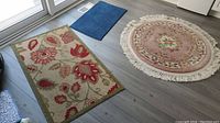 Three carpets displayed together on floor: one round beige-pink with floral design and fringe, two rectangular beige with red and green floral patterns