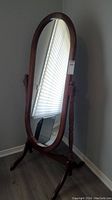 Full view of oval cheval mirror with wooden frame standing upright on wooden support frame with turned posts and gently curved feet.