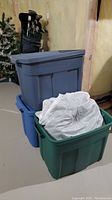 Three stacked large plastic storage bins in grey, blue, and green seen in basement setting.