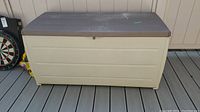 Full side view of the beige plastic deck box with dark brown lid on wooden deck, showing size and exterior condition.