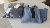 Two men's wool sweaters, one folded light blue knit with black placket detail, and one brand new sealed in plastic packaging.