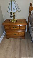 Front view of one wooden night table with two drawers, brass decorative handles, and a lamp on top (lamp not included).