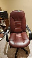 Full front view of the brown leatherette office chair showing padded seat and backrest with black plastic armrests and base.