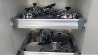 View of two kitchen drawers containing stainless steel Lagostina pots and pans with lids and black handles.