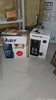 Two boxed small kitchen appliances on a concrete floor, next to a water heater: Oster bread and dough maker box and Cuisinart touchscreen coffee maker box.