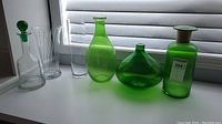 Six glass items including three green glass vases, one clear glass decanter with green stopper, and two clear glass vases, displayed on white surface near window.
