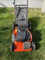 Full front view of the orange Ariens XT-7 lawnmower showing the deck, wheels, handlebar, and attached grass catcher bag on grass outdoors.