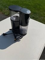 Black electric coffee grinder with clear bean hopper and ground coffee container, shown with unplugged black power cord on white surface.