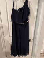Full view of navy blue formal pant suit hanging on a hanger showing asymmetrical off-the-shoulder neckline and flowy chiffon overlay.