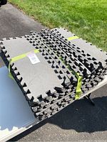 Stacked interlocking rubber gym floor tiles and edging pieces secured with yellow tape on a table outside.