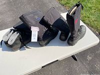 Two pairs of black men's winter boots on a white table outdoors under sunlight. One pair features the Kamik logo and adjustable buckled straps; the other is a plain black Sorel model.