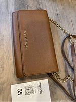 Brown Michael Kors clutch handbag lying on a wooden floor, angled showing the front flap with logo and part of the adjustable shoulder strap with gold chain.