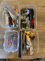 Four plastic bins showing a variety of Pokemon accessories including figurines, dice, coins, pins, and binders.