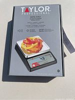 Front view of the unopened original box of the Taylor Professional digital scale showing an image of the scale with sliced bell peppers on the weighing platform and product details.