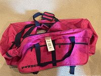 Large pink rolling duffle luggage bag with black polka dots, black straps, and external zippered compartments lying open on floor.