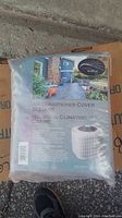 Photo of new Cover Works Air Conditioner Cover Square in original packaging showing product label and outdoor home background.