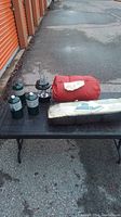 Photo of camping lot items on a black table outdoors including red sleeping bag, dome tent in carrying bag, propane tanks and lantern.