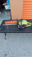 Side profile of the Poulan Wild Thing chainsaw showing green and purple body with orange bar cover on cutting bar, black handle, placed on black table.