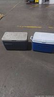 Photo showing two large coolers on a concrete floor, one dark blue with white lid (Coleman), and one gray with white lid (Rubbermaid).
