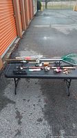 Overview photo showing three fishing poles, a fishing net, and an assortment of lures on a black folding table outside near orange storage doors.