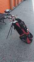 Full view of golf bag with golf clubs assembled inside, showing the black and red bag with multiple clubs inside and stand legs deployed.