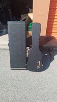 Two closed guitar cases placed outdoors, showing overall shape and size. One is rectangular, one is shaped for a guitar with Fender logo visible.