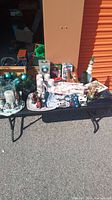Wide view of large assortment of Christmas decorations on a black folding table outdoors, including figurines, ornaments, boxes, placemats, and decorative items in green, white, and red.