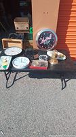 Wide shot of all clocks laid on a black table outside a storage unit including neon beer clock, wall clocks, desk clocks, mantle clock, and ceramic clock.