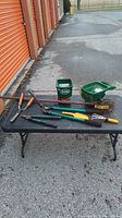 Set of garden tools laid out on a black table outside a storage unit, including hand cultivator, loppers, hoe, rake head, and seed spreaders.