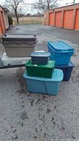 Photo showing multiple plastic storage bins stacked outdoors against storage unit doors, including green, blue, gray, and white bins.