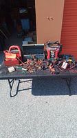 Photo showing a variety of tools spread out on table, including a red EverStart jump starter, copper fittings, clamps, toolbox, power tools, and electrical testers